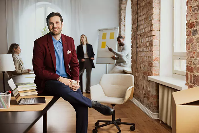 Hyväntuulinen mies istuu toimistopöydällä. Taustalla ihmiset häärivät fläppitaulun ympärillä. 